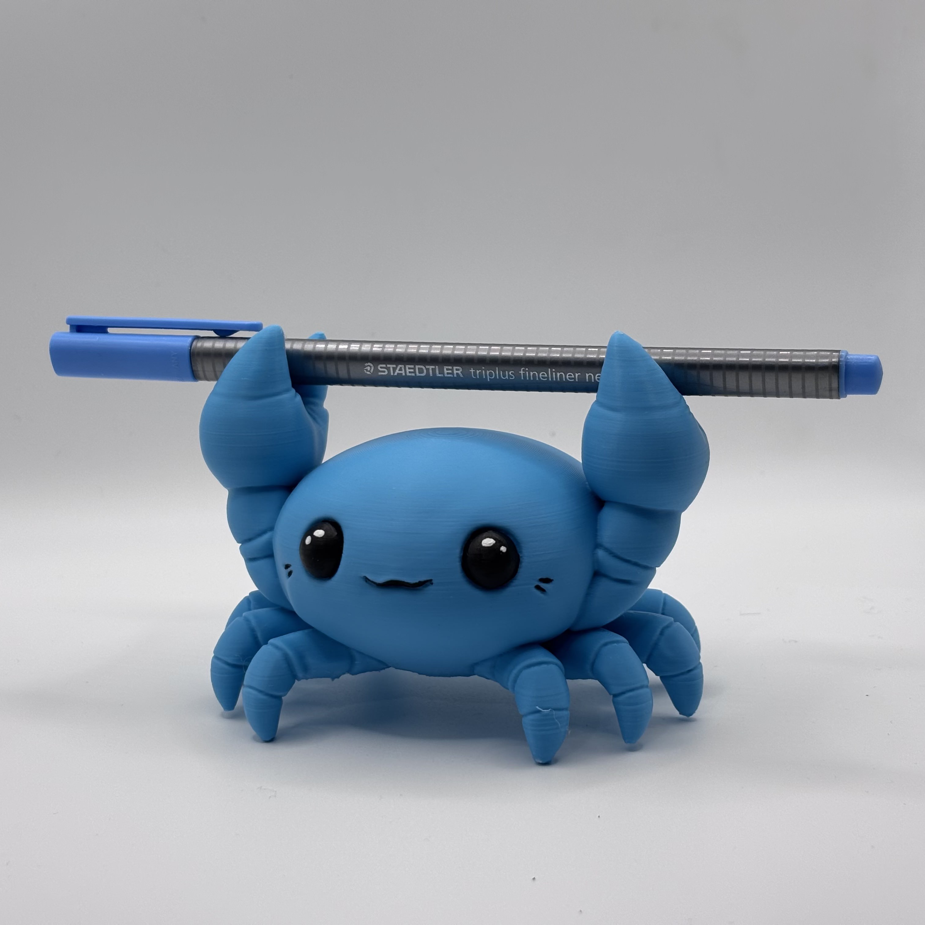 Blue chibi crab pen holder 3D printed in PLA, holding a Staedtler fineliner pen as a functional desk accessory.